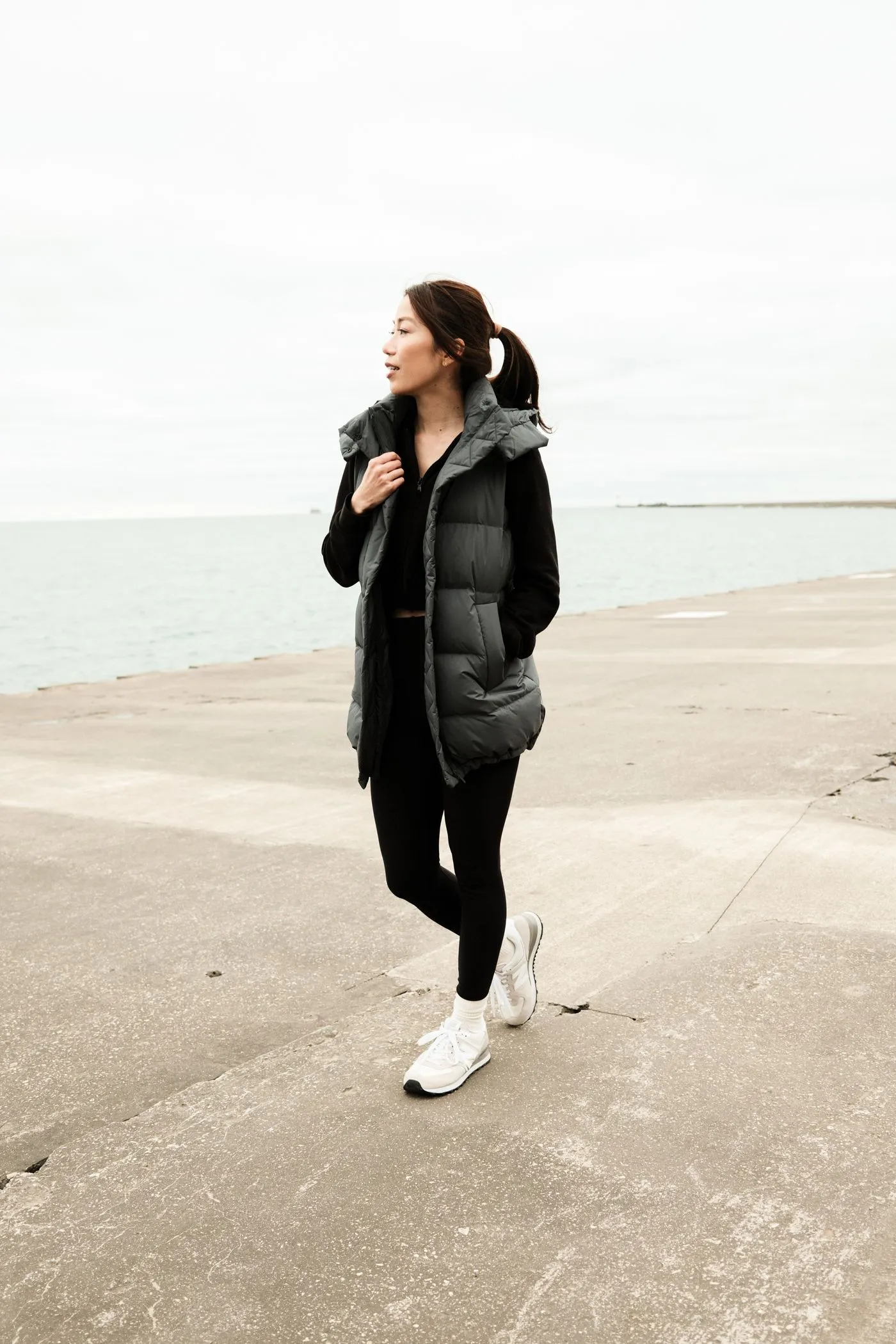 A woman in a long gray puffer vest walking along a lakeside path on a cold, gray day